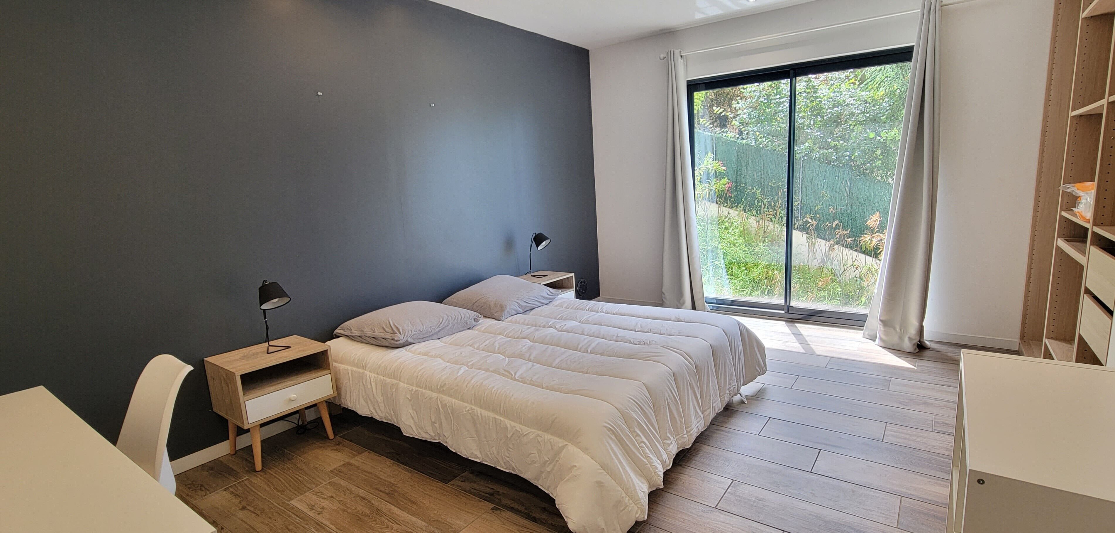 A bedroom with a large bed, a bedside table, a lamp, and a view of greenery through a window.