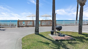 Un chemin pavé mène à une zone de plage agrémentée de palmiers, d’un barbecue et de parasols.