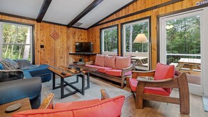 A cozy cabin interior with a wooden interior, a sofa, chairs, a coffee table, and large windows offering a view of the forest.