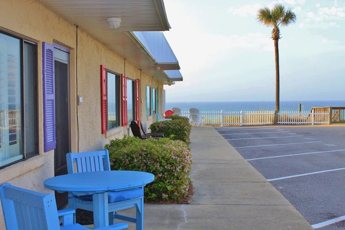 A motel with a parking lot, a table and chairs outside, and a view of the ocean.