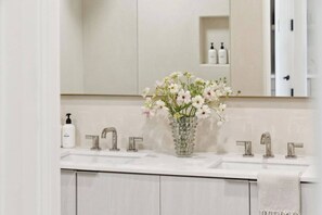 A bathroom with a white sink, three faucets, a glass vase with flowers, and a mirror above the sink.