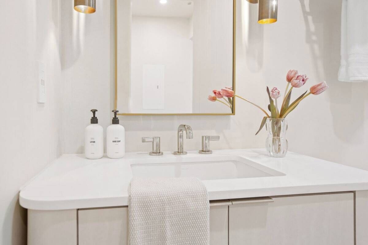 Un baño moderno con un lavabo blanco, dos dispensadores de jabón para manos, un espejo con marco dorado y un jarrón con flores rosadas.