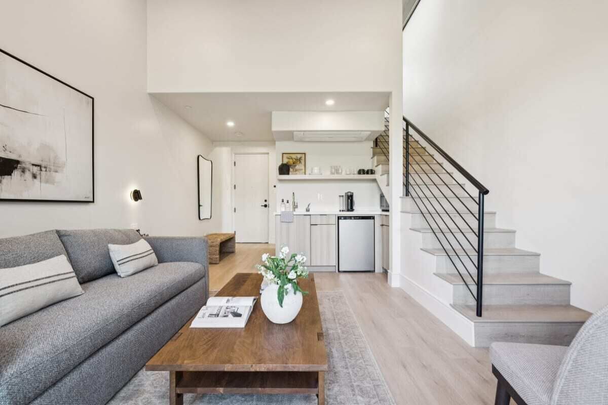 A modern living room with a grey sofa, a wooden coffee table with a vase of flowers, and a staircase with a black railing leading to a kitchen area.