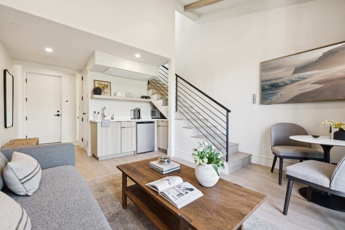 Un salon moderne avec un canapé, une table basse et un coin repas avec des chaises. On aperçoit une cuisine au fond, avec un escalier menant à un autre niveau.