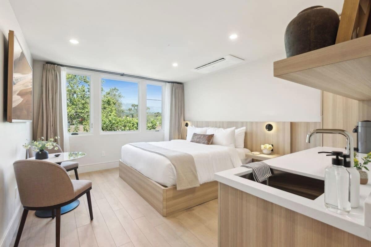 A modern bedroom with a large bed, bedside tables, a desk, and a window with a view of greenery.