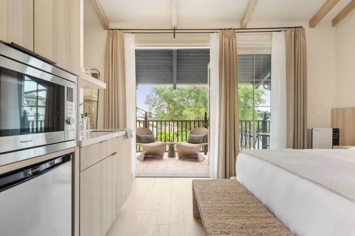 Habitación de hotel compacta con kitchenette, una cama, un banco y un balcón con sillas.