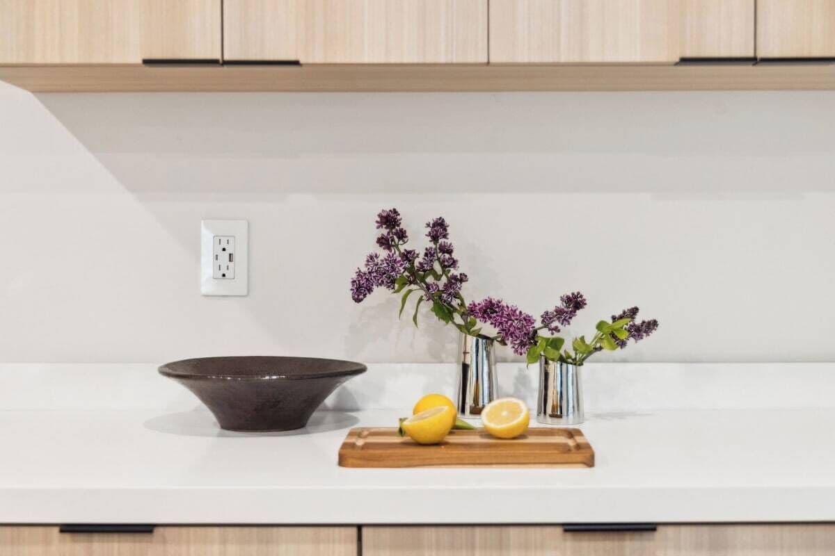 Una encimera de cocina con un bol oscuro, una tabla de cortar de madera, dos limones y un florero con flores moradas.