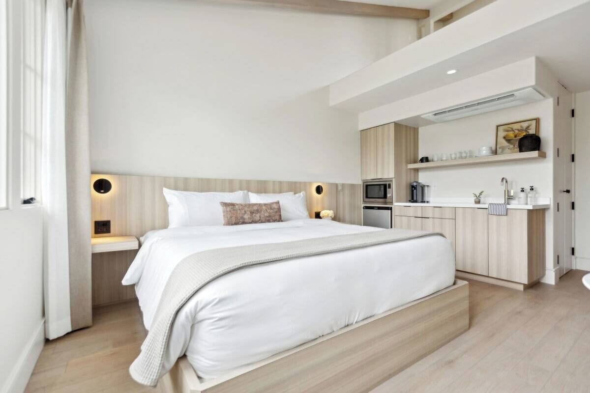 A modern bedroom with a large bed, bedside tables, a built-in bench, a kitchenette, and a bathroom.
