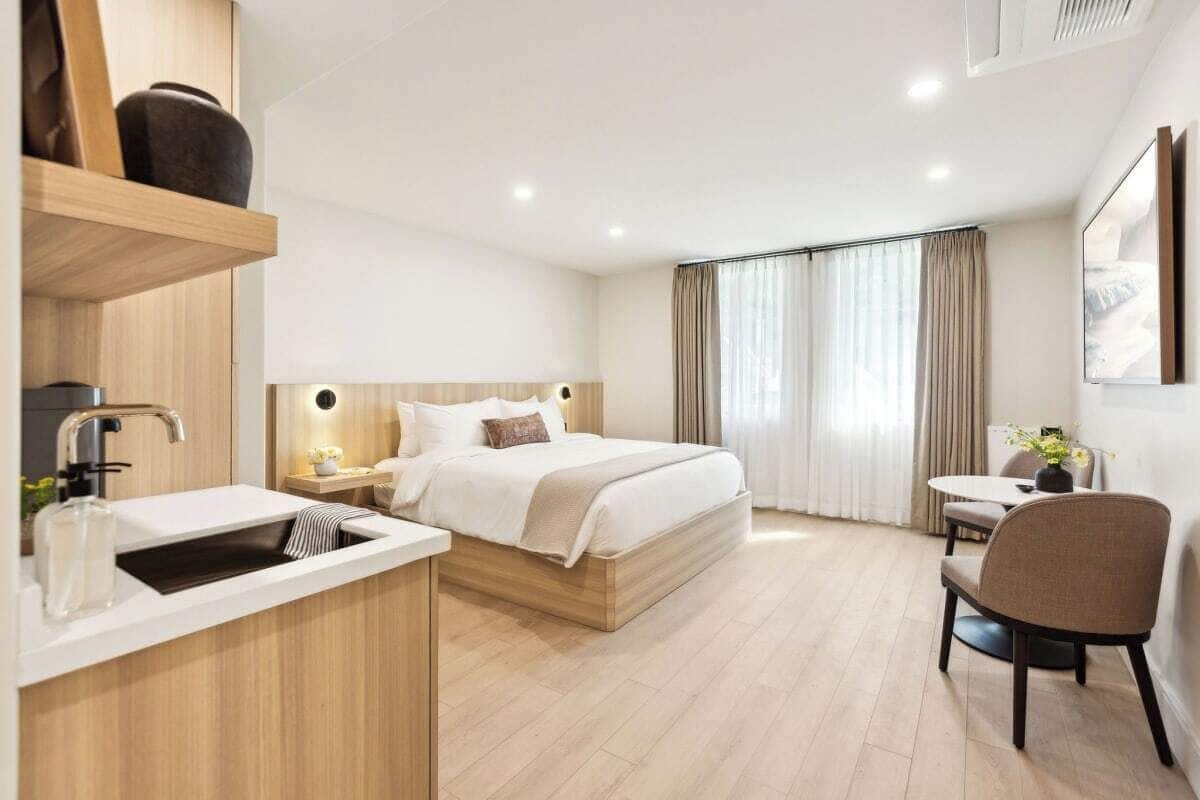 A modern hotel room with a wooden bed, a small desk, a chair, and a large window with curtains.