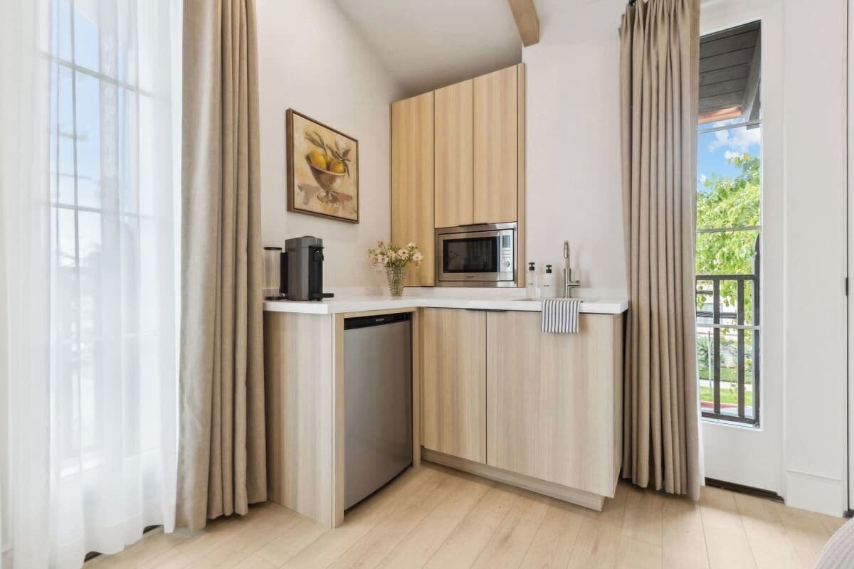 Una cocina funcional equipada con microondas, cafetera y lavavajillas. Cuenta con muebles de madera clara, una pequeña encimera y vistas al exterior con vegetación.