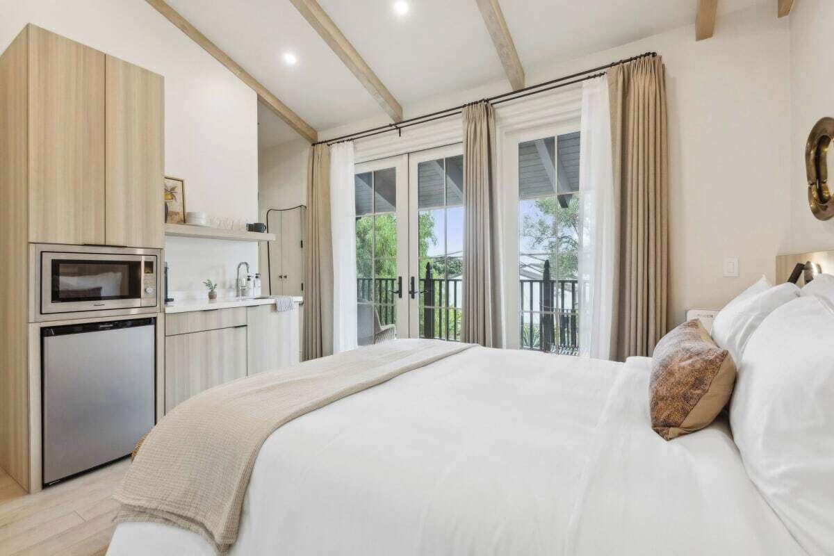A bedroom with a bed, a small kitchenette, and a balcony with curtains.