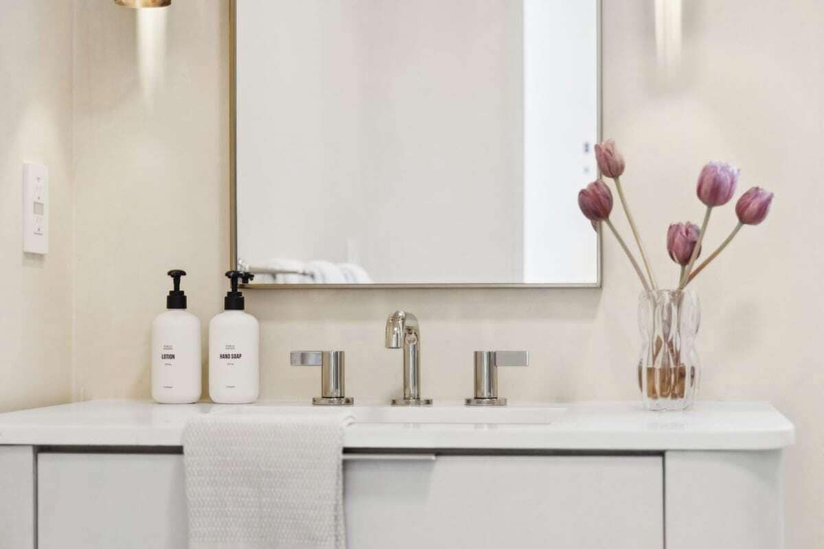 A bathroom vanity with a mirror, two bottles of lotion, a faucet, and a vase with flowers.