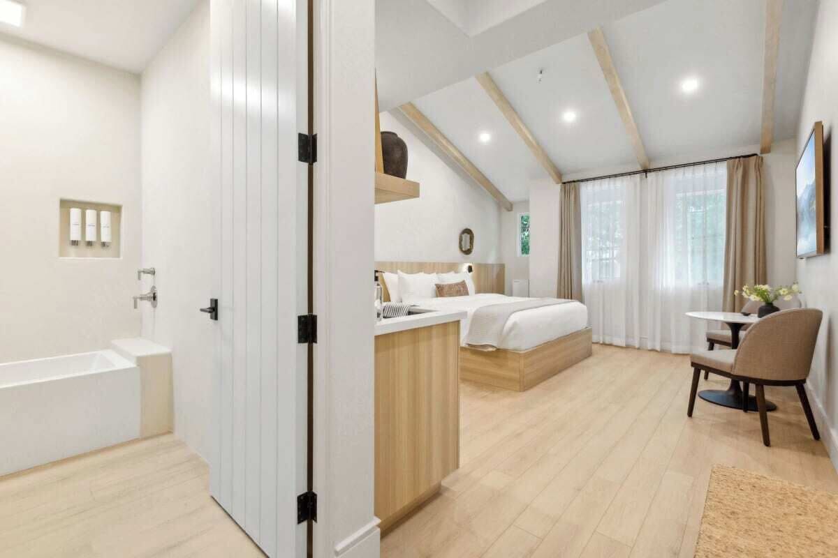 A modern hotel room with a large bed, a separate sitting area, a small kitchenette, and a bathroom with a bathtub.