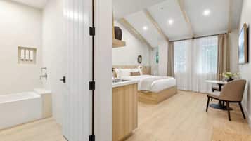 A modern hotel room with a large bed, a separate sitting area, a small kitchenette, and a bathroom with a bathtub.