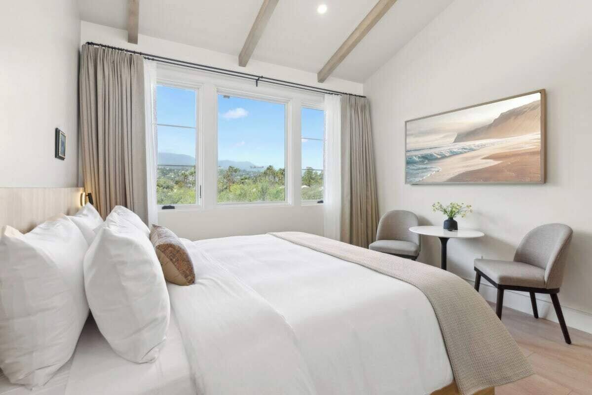 A bedroom with a large bed, a view of the outdoors, a small table with a chair, and a painting on the wall.