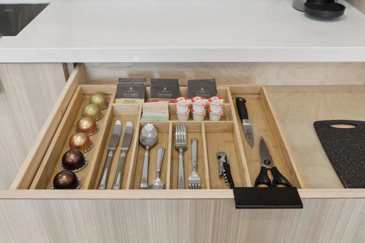Un cajón con cubiertos ordenados, incluyendo cucharas, tenedores y cuchillos, junto con recipientes pequeños y un par de tijeras.