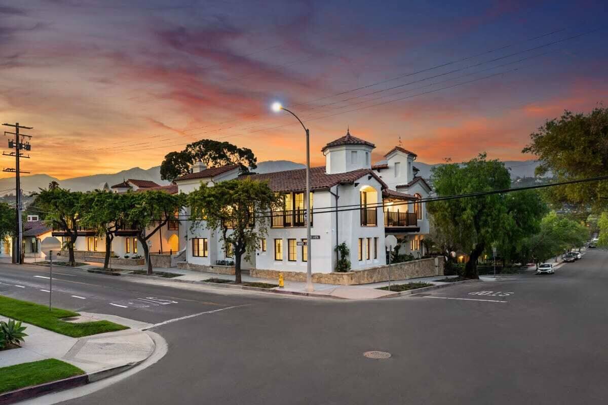 A two-story building with a white facade and a red-tiled roof, situated at a street corner with a clear sky and sunset.