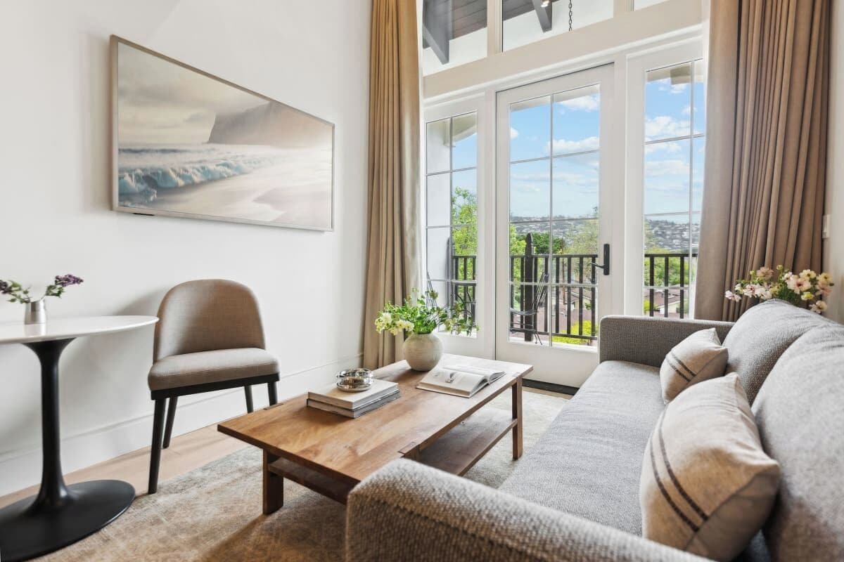 A modern living room with a sofa, a coffee table, a chair, and a painting on the wall. There is a balcony with a view of the city.