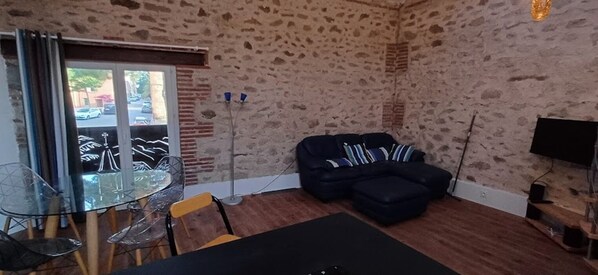 Uma sala de estar com parede de pedra, uma mesa de vidro com cadeiras, um sofá preto e uma TV na parede.