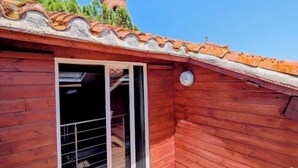 Une terrasse en bois surmontée d’un toit recouvert de tuiles rouges, une porte-fenêtre coulissante et, au fond, un petit clocher.