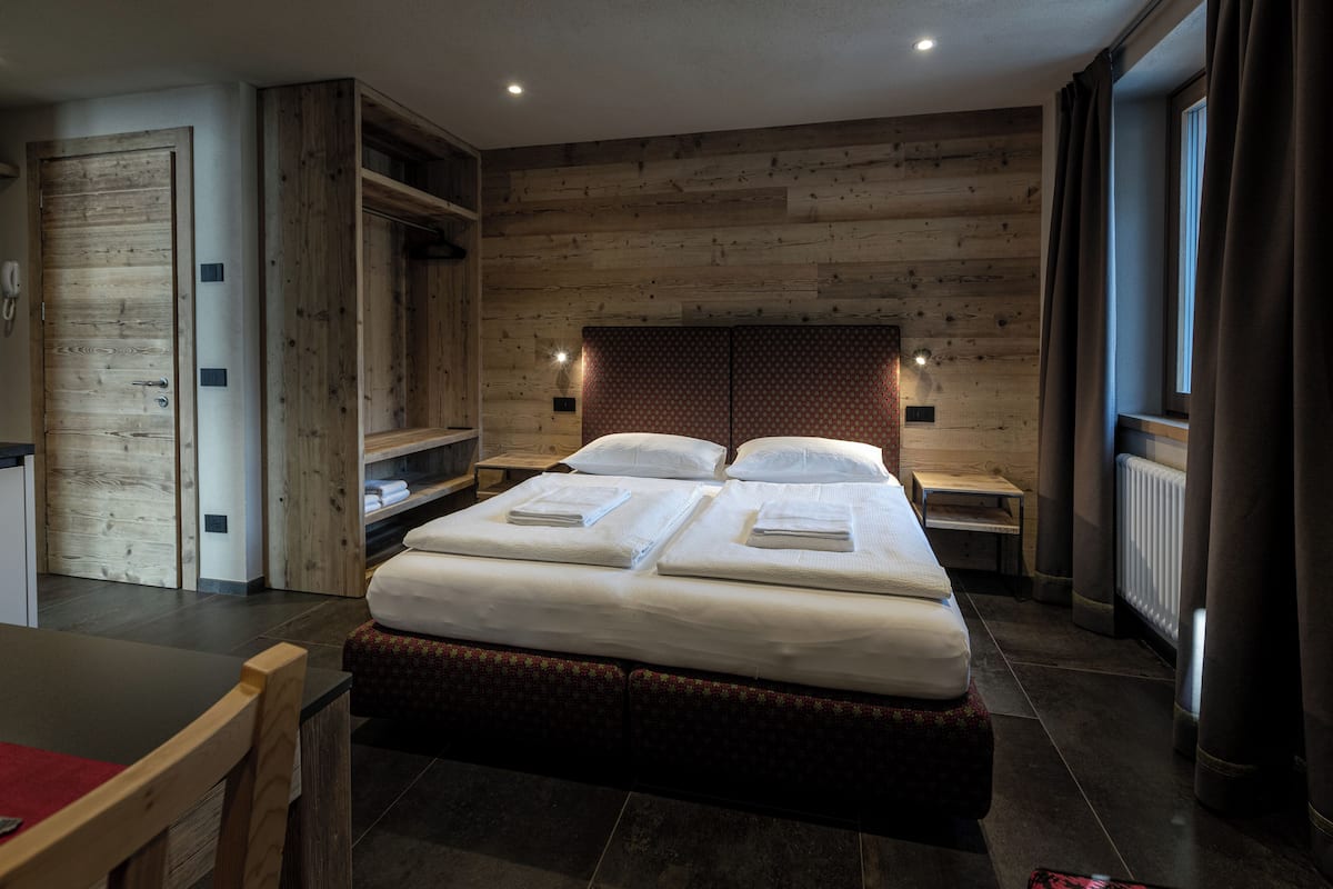 Quarto de hotel com parede de madeira, uma cama grande com duas almofadas, mesa de cabeceira, uma mesinha e uma cadeira.