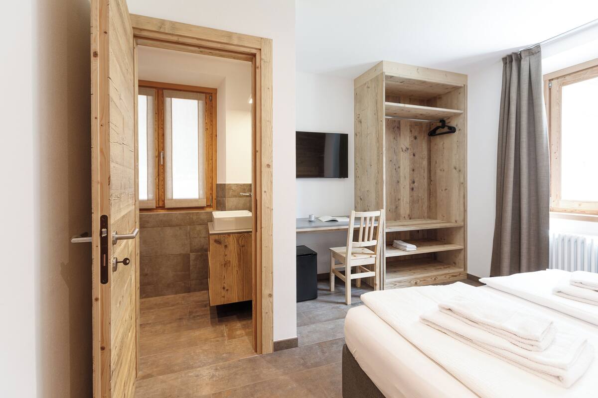 Quarto de hotel com guarda-roupa de madeira, escrivaninha, cadeira, cama e televisão.