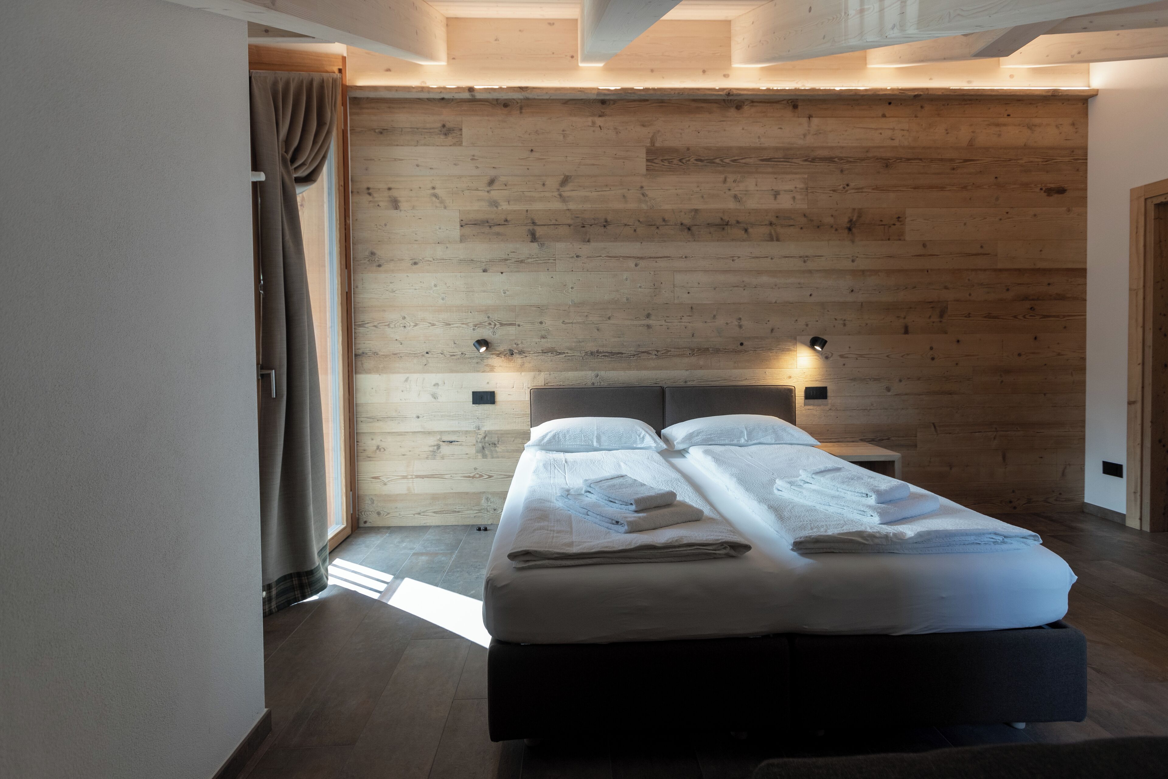 A modern bedroom with a wooden wall, two beds, bedside lamps, and a large window.