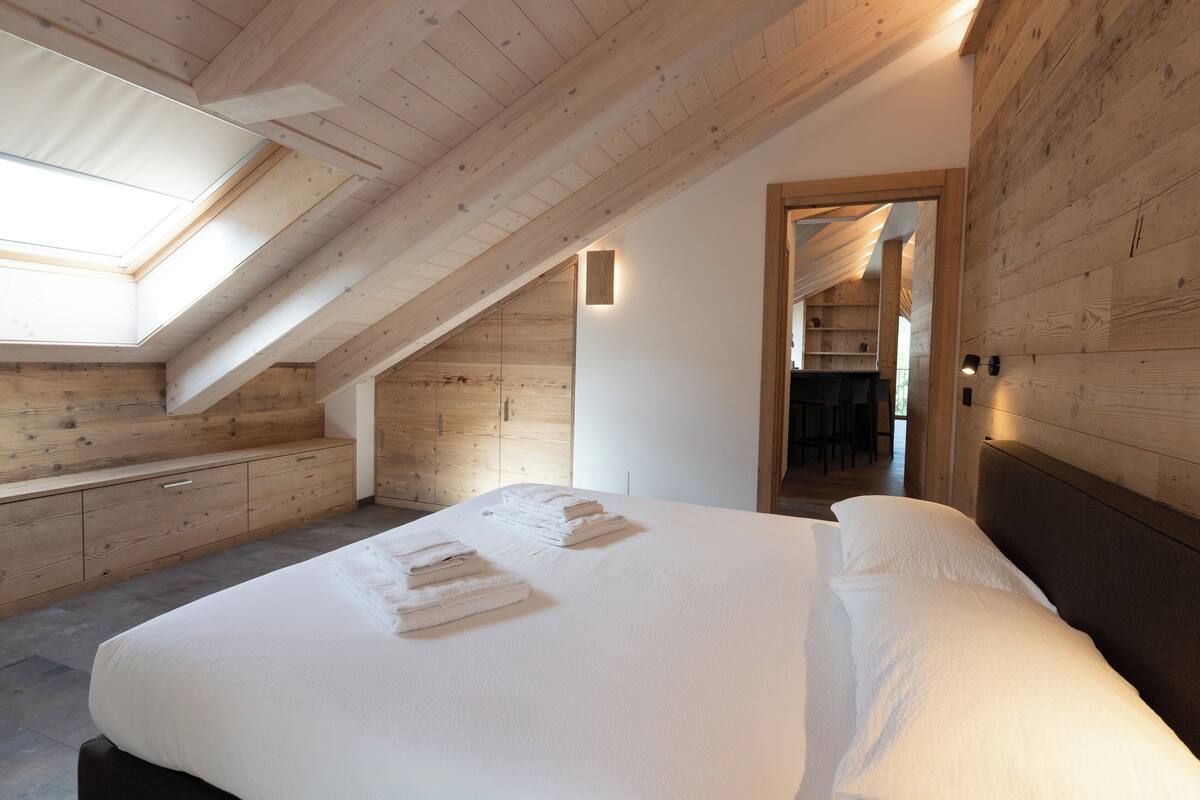 Quarto com claraboia, paredes de madeira, cama com roupa de cama branca e um banco embutido.