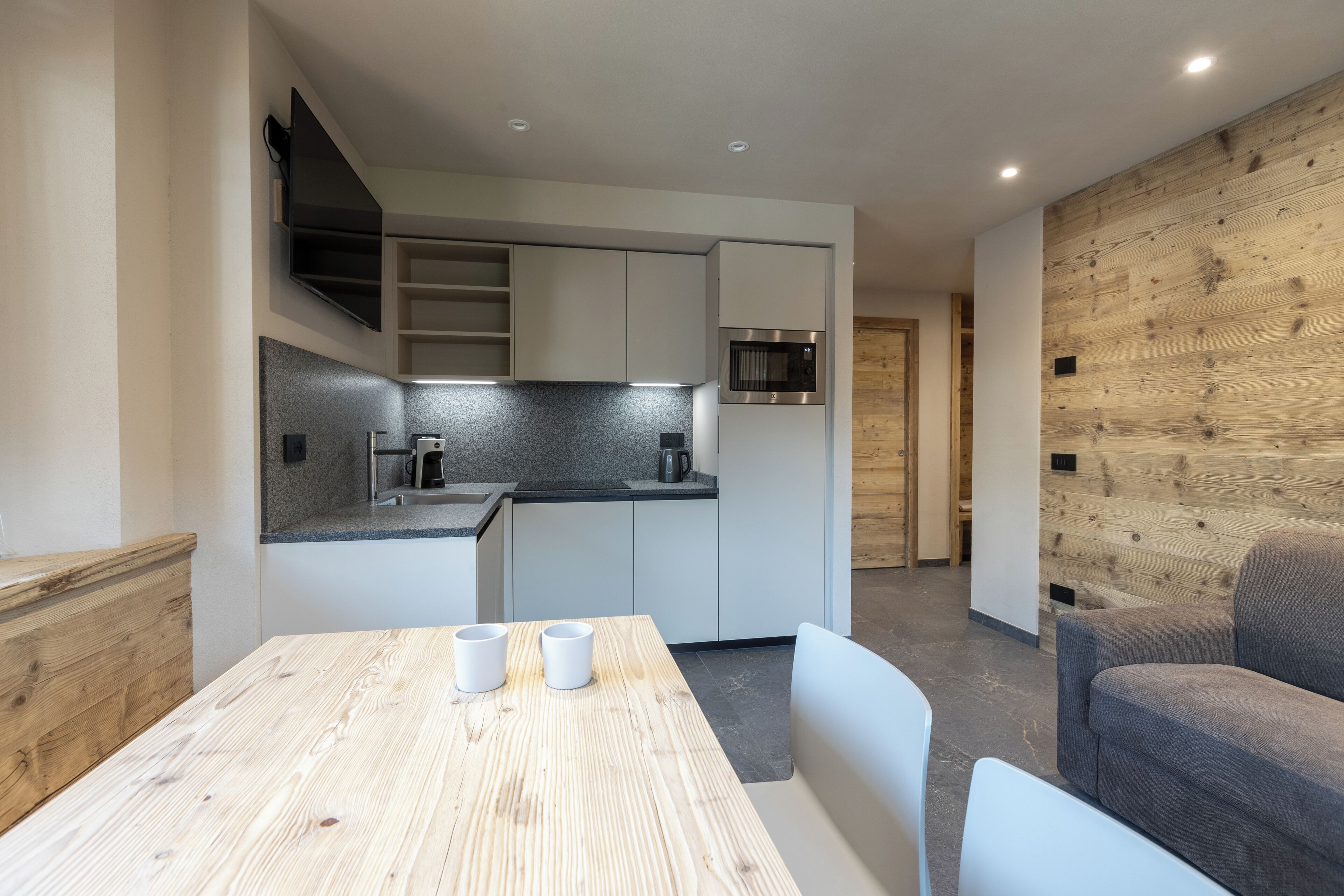 Une cuisine moderne avec des meubles blancs, un four à micro-ondes encastré et un mur en bois. On y trouve une table à manger avec deux chaises blanches et un canapé gris.