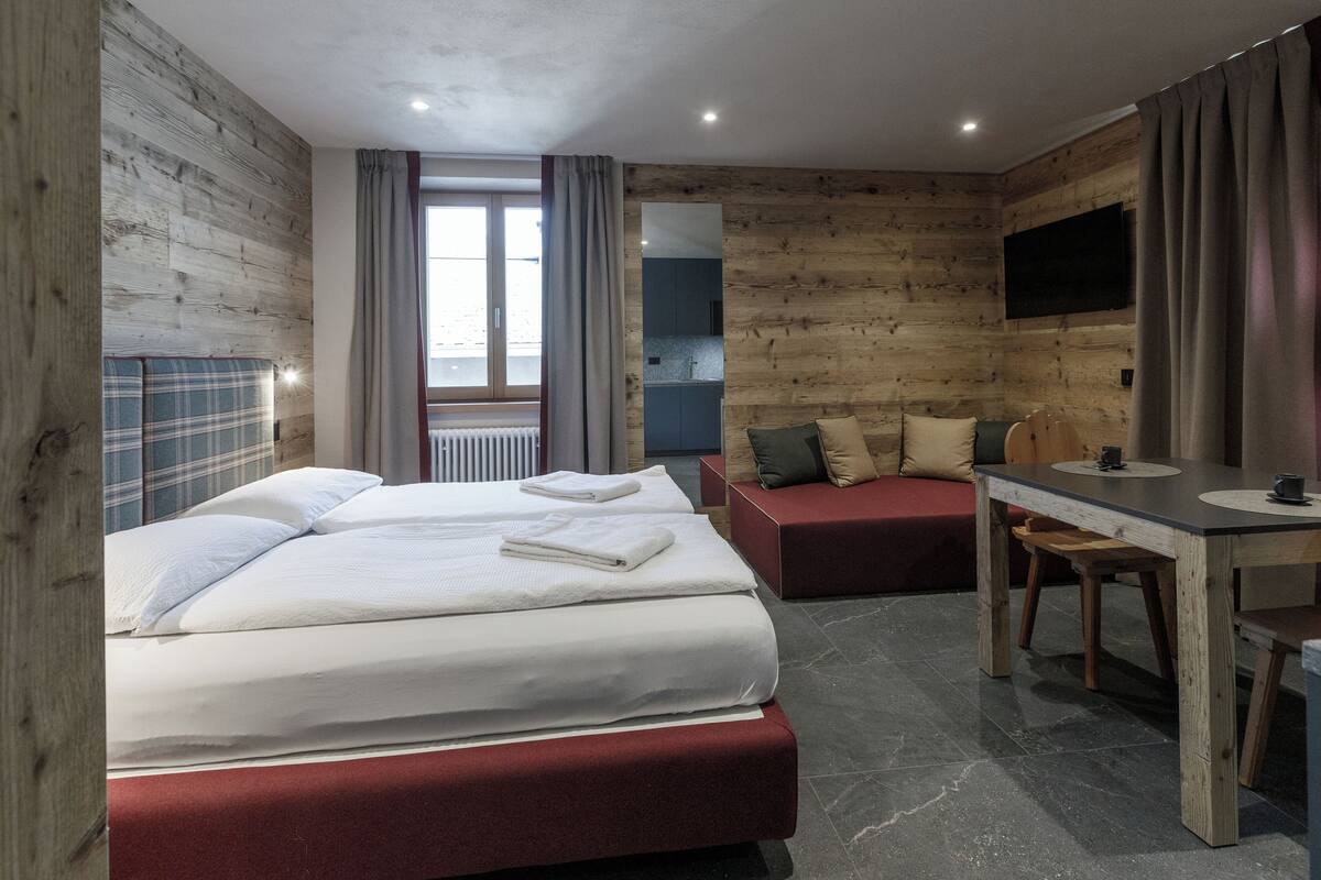 Quarto de hotel com parede de madeira, cama com roupa de cama branca, sofá vermelho, uma mesinha com uma chávena e uma televisão na parede.