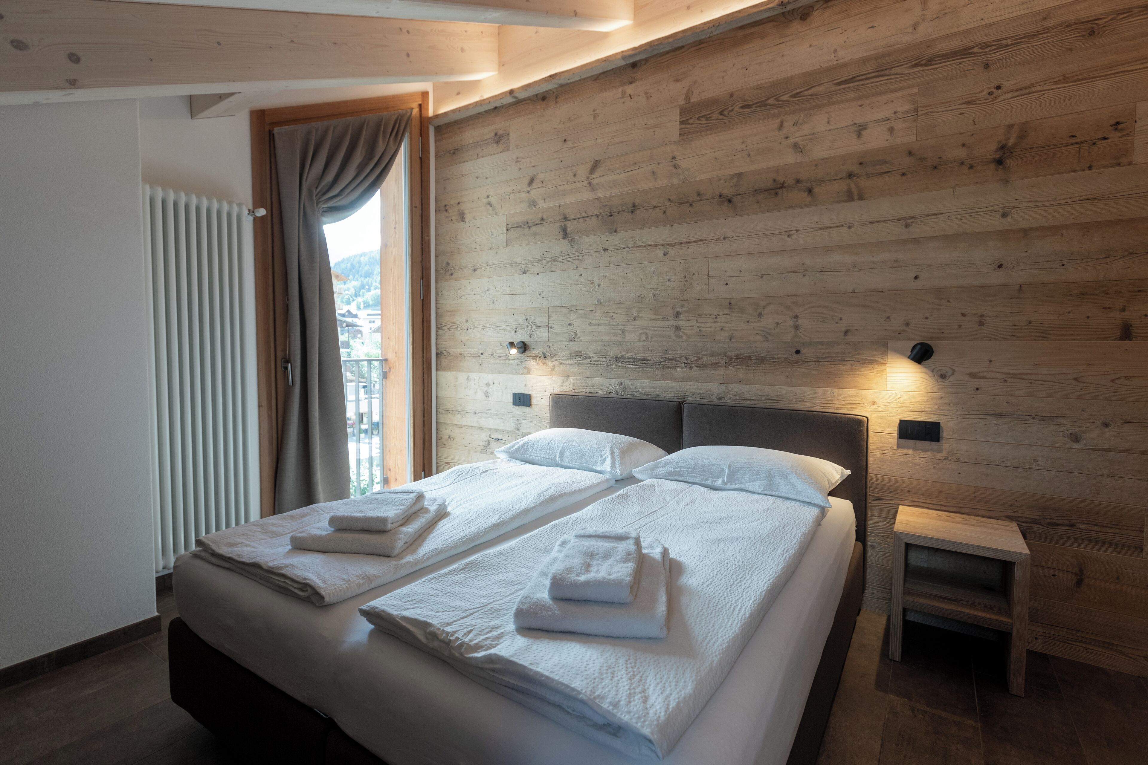 Cama doble con ropa de cama y toallas blancas, cabecera de madera, mesita de noche y ventana con cortinas.
