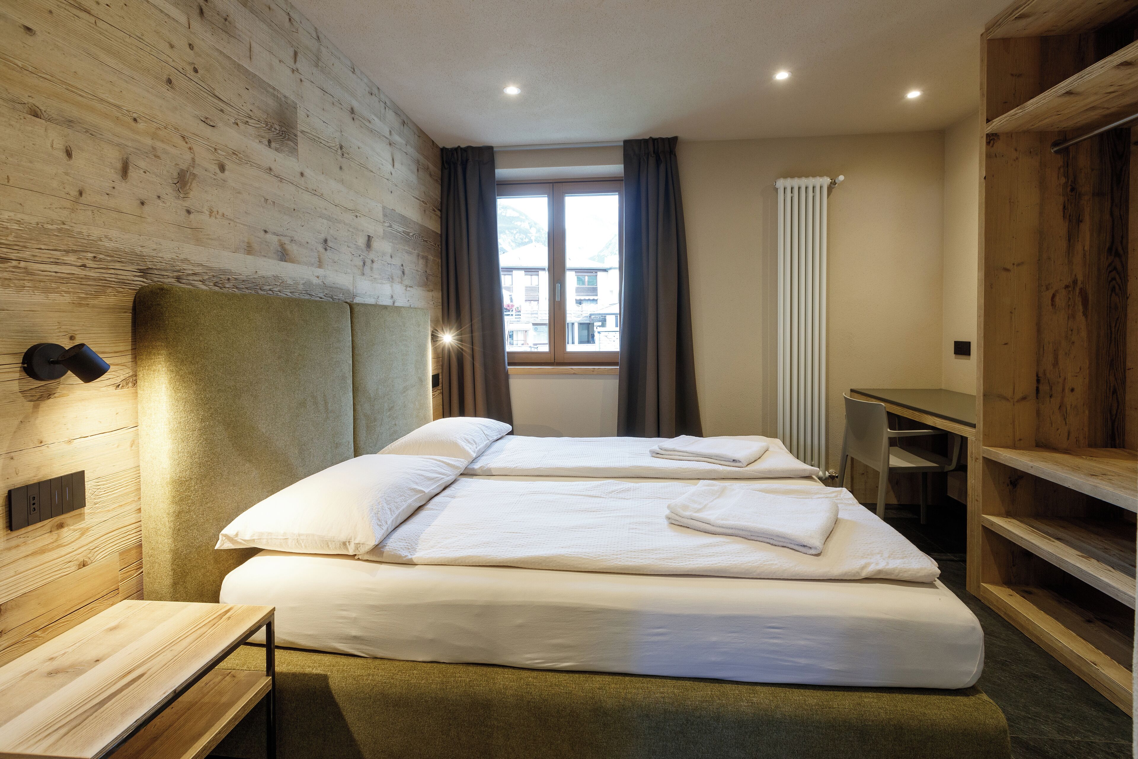Ein Doppelbett mit weißer Bettwäsche, ein hölzerner Kopfseitenverzierer, ein Nachttisch, ein Fenster mit Vorhängen und eine Heizung.