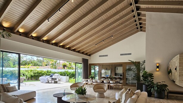 A spacious living area with a high ceiling, built-in wooden beams, a large sofa set, a coffee table, and a kitchen area visible through an open space.