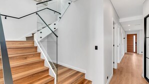 Un escalier moderne avec des rampes en verre et des marches en bois, menant à un couloir doté d’un plancher en bois et de murs blancs.