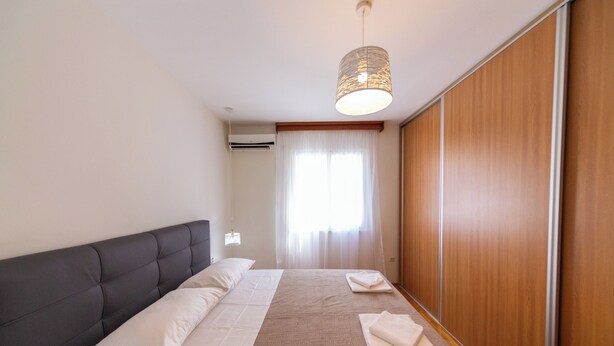 A hotel room with a bed, a bedside table, a hanging light fixture, a window with curtains, and a wooden wardrobe. - Lovely 2 Bdr Apartment in Heart of Kotor (Dobrota)