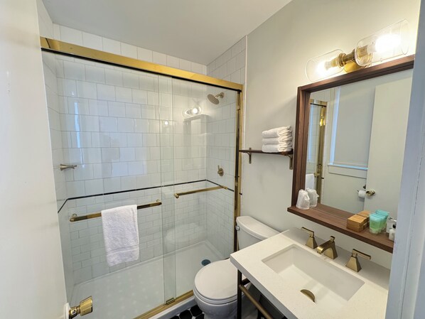A modern bathroom with a glass-enclosed shower, white tiled walls, a white sink with gold fixtures, and a large mirror with a wooden frame.
