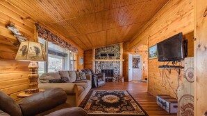 A cozy cabin interior with a stone fireplace, leather sofas, a television, and a wooden ceiling.