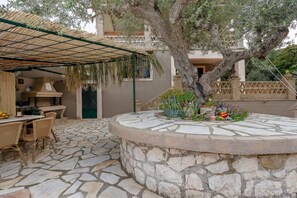 A stone-floored patio with a round stone table and wicker chairs, a pergola with hanging plants, and a large tree in the center. - Vadina Apartment 1 in Zakynthos (Zakynthos)