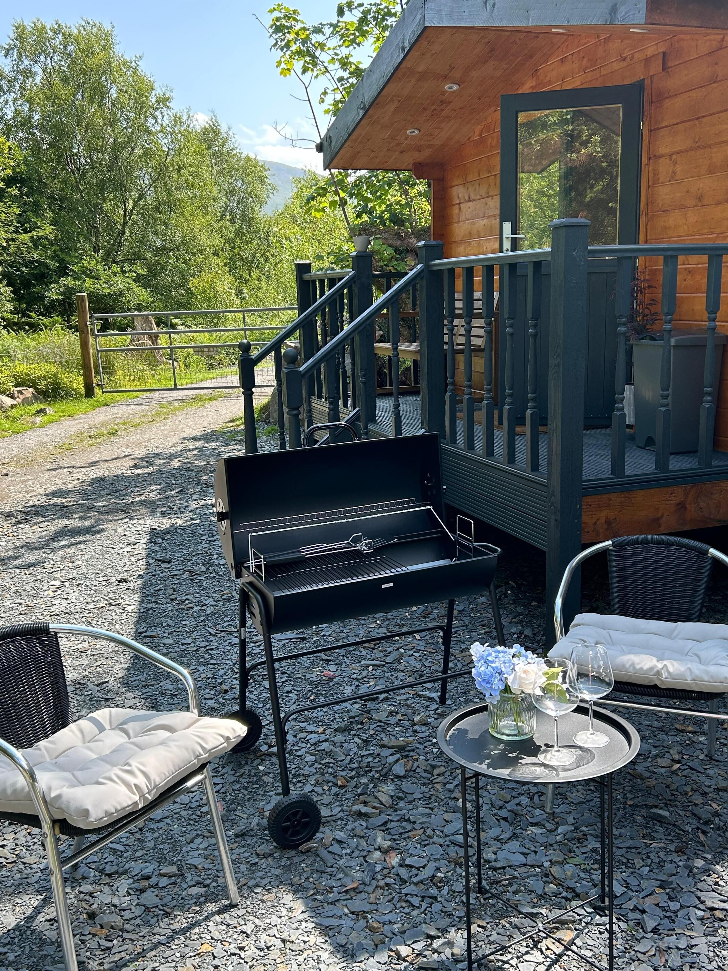 A wooden cabin with a balcony, a metal grill, outdoor seating, and a gravel path.