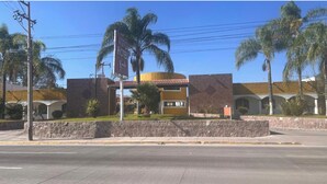 Un hotel de dos pisos, con una entrada central y un jardín delantero arbolado con palmeras.
