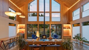A spacious room with wooden walls, large windows, a central seating area with a fire feature, and potted plants.