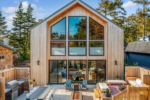 Una cabaña moderna de madera con ventanas amplias, un patio con mobiliario exterior y una fogata.