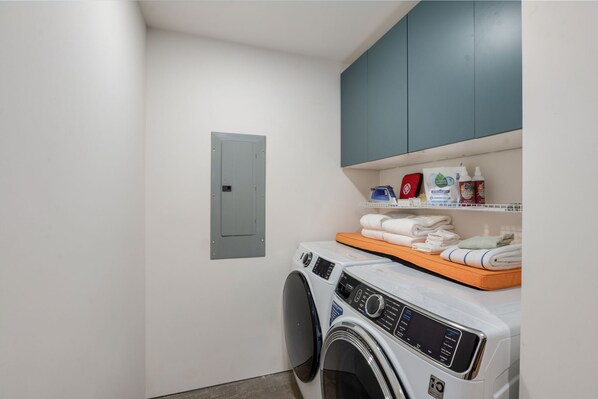 Un lavadero con lavadora, secadora, un estante con productos de limpieza y una puerta gris.