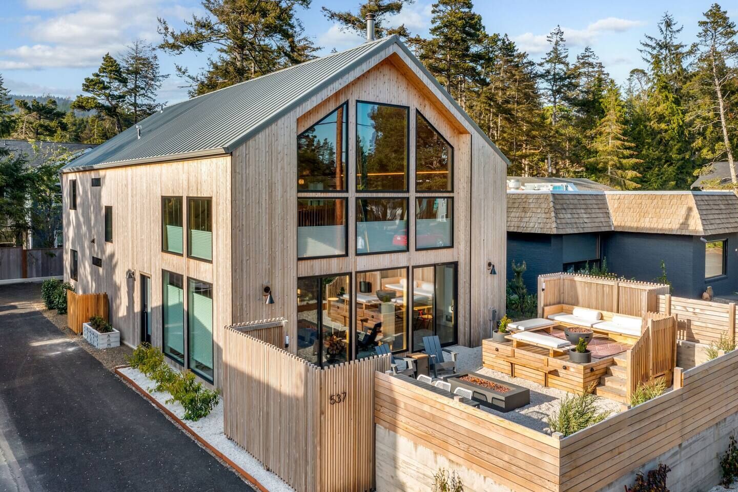 A modern wooden cabin with large windows, a deck with seating, and a view of the surrounding forest.