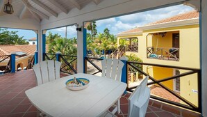Un espace extérieur abrité, avec une table et des chaises blanches, un saladier rempli de fruits et une vue sur un bâtiment jaune doté d’un balcon.