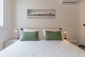 A neatly made bed with white linens and green pillows, a cityscape painting on the wall, and a window on the left.