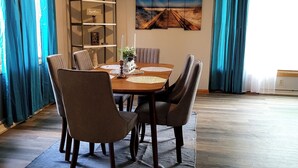 A dining area with a wooden table, chairs, and a rug. There is a shelf with decorative items and a painting on the wall.