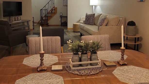 A dining area with a wooden table, chairs, and a chandelier. There is a sofa and a television in the background.