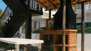 An outdoor patio with a brick barbecue grill, a white table, and metal chairs. - Freedom Surf House (Peniche)