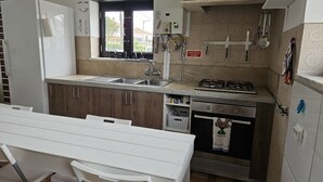 A compact kitchen with a sink, stove, and oven. There's a window above the sink and a sign indicating a gas cut-off point. - Freedom Surf House (Peniche)